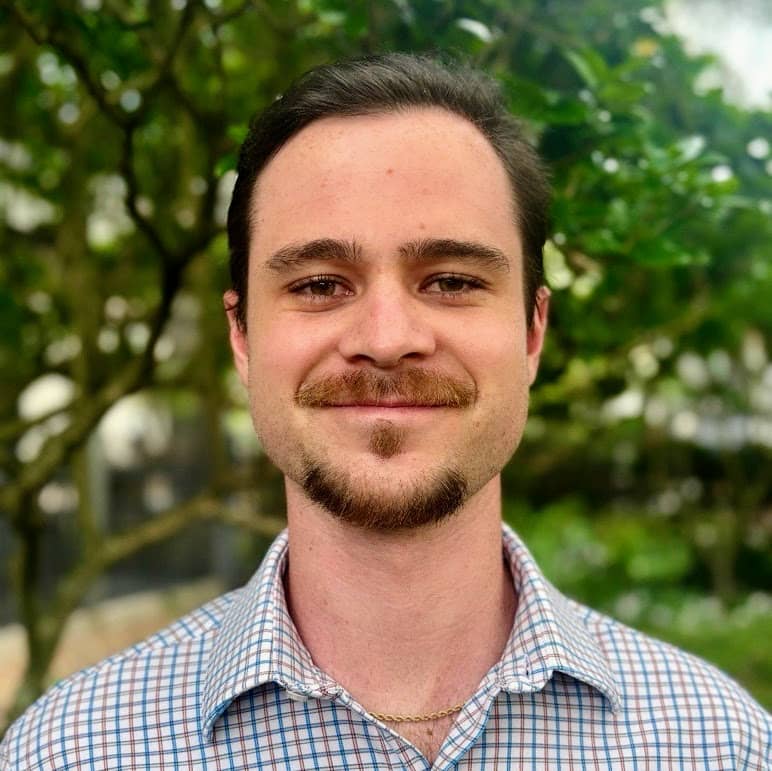 Professional headshot of Brendan Walsh, smiling outdoors with a natural green leafy background, wearing a light checkered shirt.