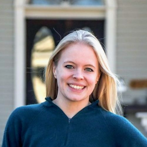 Caroline Boyd-Rogers, a woman with long blonde hair wearing a dark green sweater, smiles in front of a house with a wooden door and windows in the background.