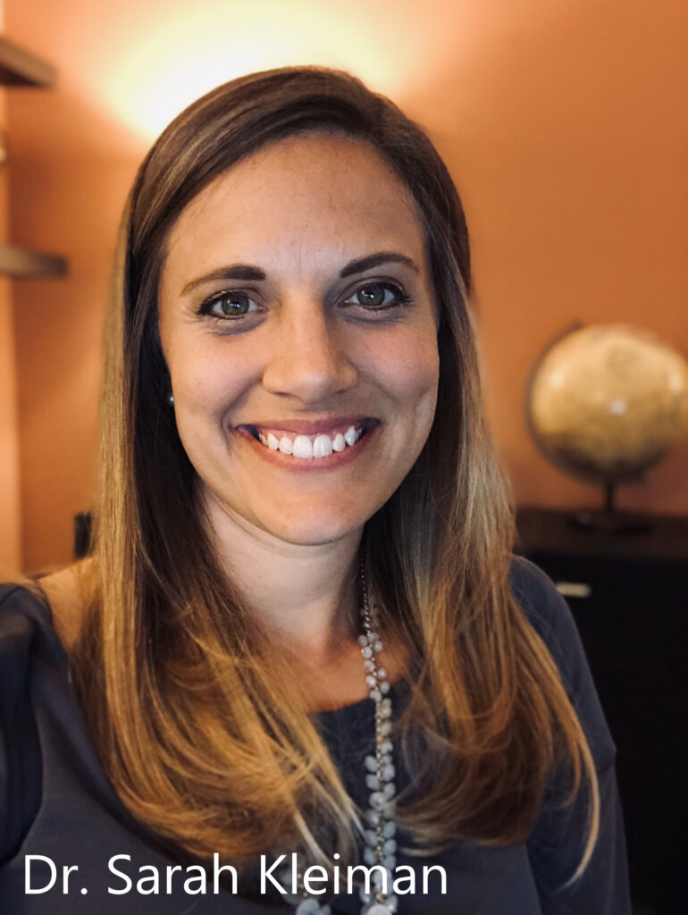 A woman with long blonde hair smiling warmly in a softly lit office with a globe in the background. The text "Dr. Sarah Kleiman" is overlaid in white at the bottom.