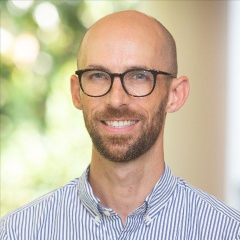 Jeremy Eberle smiling, wearing glasses and a striped shirt, in a professional portrait with a blurred natural background.