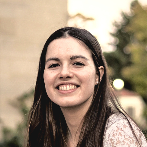 Linnea Sepe-Forrest, a young woman with long dark brown hair, smiling outdoors with a blurred background of trees and buildings.