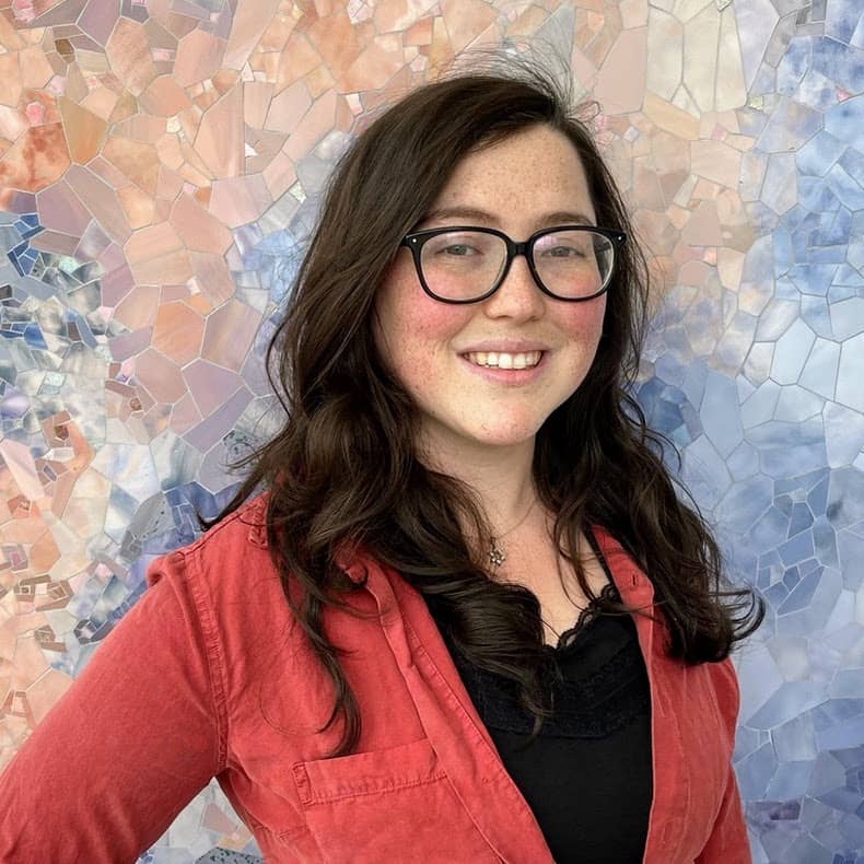 Headshot of Rachel Telles, a graduate student from the University of Iowa, smiling outdoors with green foliage in the background.