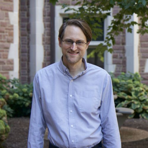 Tom Rodebaugh, Ph.D., smiling outdoors in front of a brick building with greenery in the background.